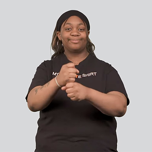 The image shows a person standing against a plain background. The person is wearing a black shirt with text on it. The person is making the sign for coffee. 