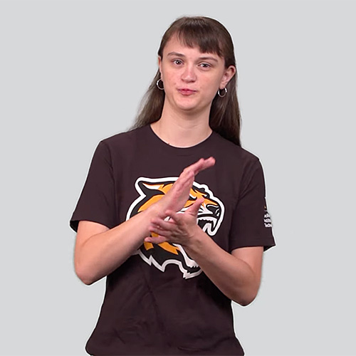 A person with long brown hair and hoop earrings, wearing a brown T-shirt featuring a tiger mascot logo, signs in ASL against a solid grey background.