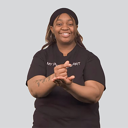 The image shows a person standing against a plain background. They are wearing a black t-shirt. They are making the sign for graduation.
