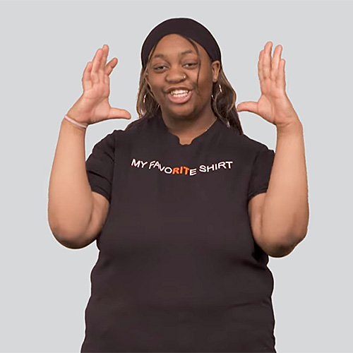 The image shows a person standing against a plain gray background. The person is wearing a black t-shirt. They are making the sign for imagination.
