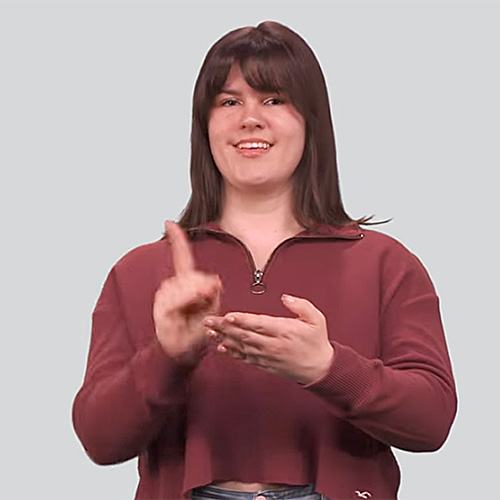 The image features a young woman with shoulder-length dark brown hair, wearing a maroon cropped sweater. She is smiling and using sign language. The background is a plain, light gray color.