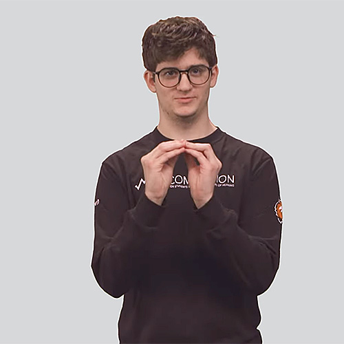 The image features a young man with short brown hair, glasses, and light skin, wearing a black long-sleeve shirt and khaki pants. He is making a hand gesture in front of his chest, where the tips of his fingers from both hands touch, forming the sign ‘addition.’ His facial expression is neutral with a slight smile. The background is a solid light gray color.