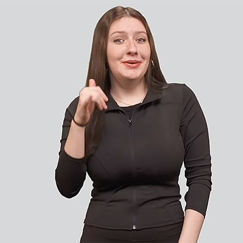 The image features a woman dressed in black, making a hand gesture in American Sign Language (ASL). She has long brown hair and is smiling while looking towards the camera. The background is a plain, light gray color.