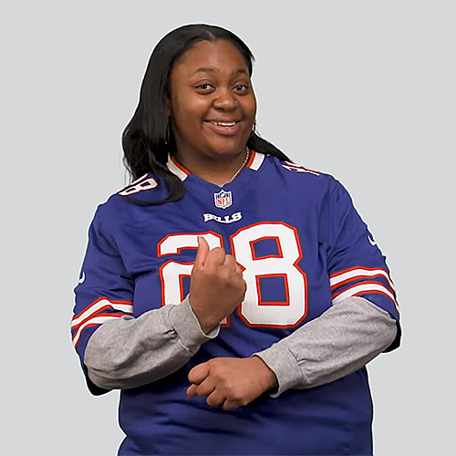 The image features a woman wearing a blue Buffalo Bills football jersey with the number 28 on it. She has a friendly and confident expression with a smile on her face. She is making a sign with her hands in American Sign Language (ASL) for the word "Win." She is wearing a gray long-sleeve shirt underneath her jersey. The background is a plain light gray color.
