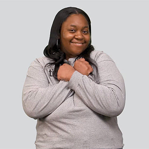 The image features a woman with dark hair, smiling and crossing her arms over her chest, making the ASL sign for love. She is wearing a light gray long-sleeve shirt. The background is a plain light gray color.