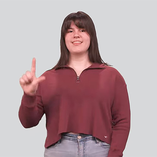 The image shows a person with shoulder-length dark hair wearing a maroon long-sleeve shirt and jeans. They are making the American Sign Language (ASL) sign for "Library" by forming an "L" shape with their right hand and rotating it in a circular motion. The background is a plain light color.