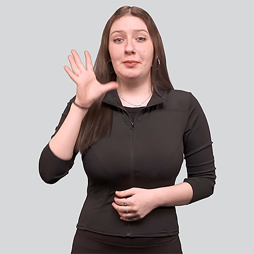 The image features a woman dressed in black, making a hand gesture in American Sign Language (ASL). She has long brown hair and a neutral facial expression while looking towards the camera. The background is a plain, light gray color.