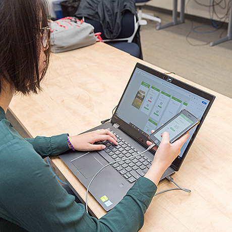 A person is sitting at a wooden desk using a laptop and holding a smartphone. The laptop screen displays a mobile app design interface with various screens laid out, suggesting they are working on app development or testing. A charging cable connects the phone to the laptop.