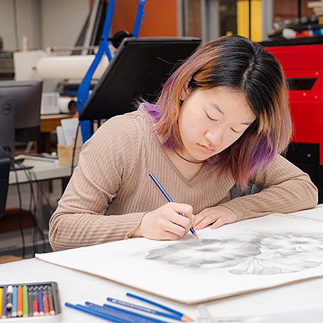**Alt text:** A person with shoulder-length hair, partially dyed purple, is seated at a desk intently drawing a detailed flower sketch with a colored pencil. Various colored pencils are scattered on the table, and the background includes art studio equipment and furniture.