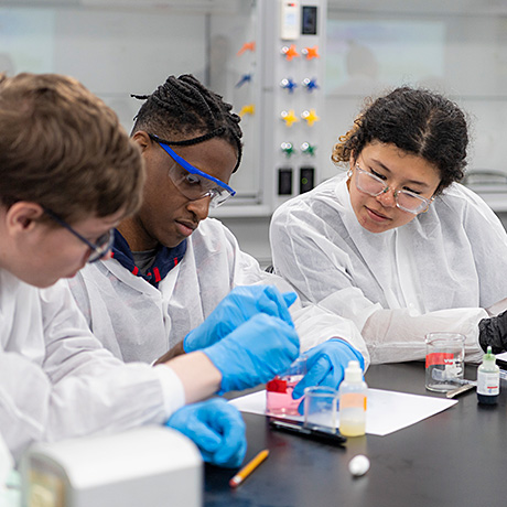 **Alt text:** Three students wearing white lab coats, safety goggles, and blue gloves are working together at a lab bench. They are engaged in a hands-on science experiment, using beakers with colorful liquids and dropper bottles, in a laboratory setting.