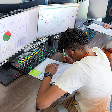 Person sitting at a desk with multiple computer monitors displaying financial charts and graphs, writing on a piece of paper.