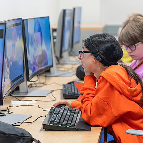 Students sitting at a row of computer desks, focused on their screens. One student in an orange hoodie is prominently visible, with others blurred in the background.