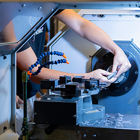 Person operating a CNC machine, adjusting a workpiece inside the machine with visible metal shavings and coolant hoses.