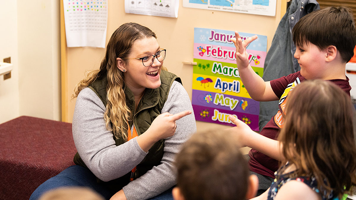 A teacher and children in a classroom using sign language. The background shows colorful posters with the names of months.