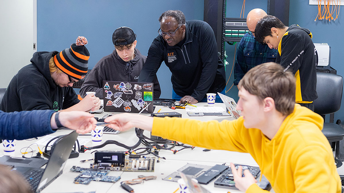 Students seated around a table work on computer hardware while a professor looks on