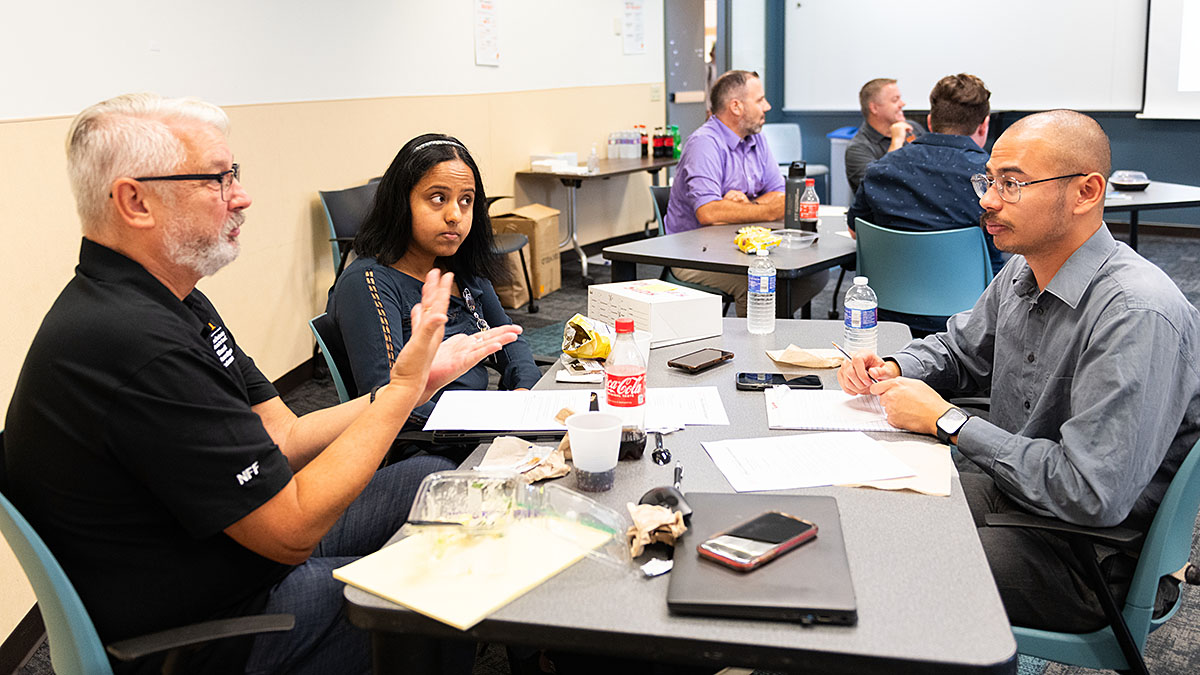 Group of people sitting around tables in a classroom setting, engaged in discussion and working on papers.