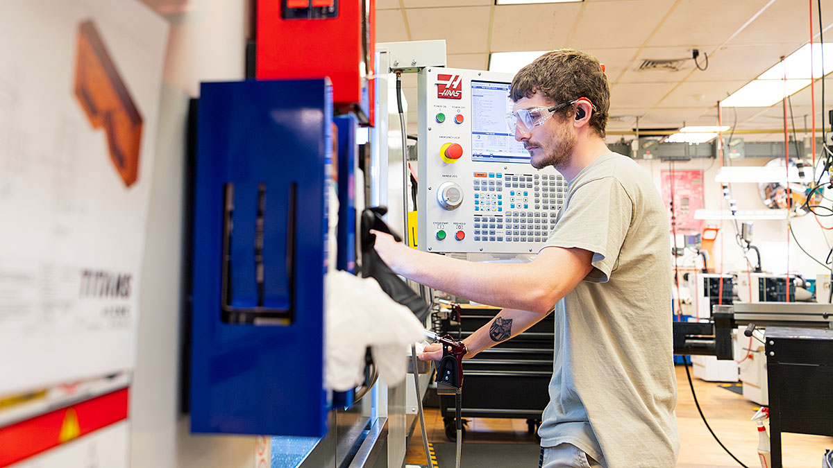 Person operating a Haas CNC machine in a workshop, wearing safety glasses and ear protection, focused on the control panel in an industrial setting.