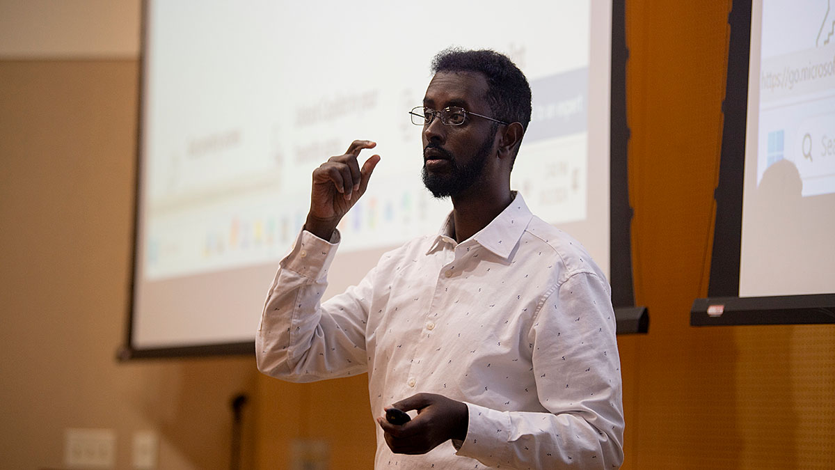 Person giving a presentation with two projection screens in the background, wearing a white shirt and holding a remote control.