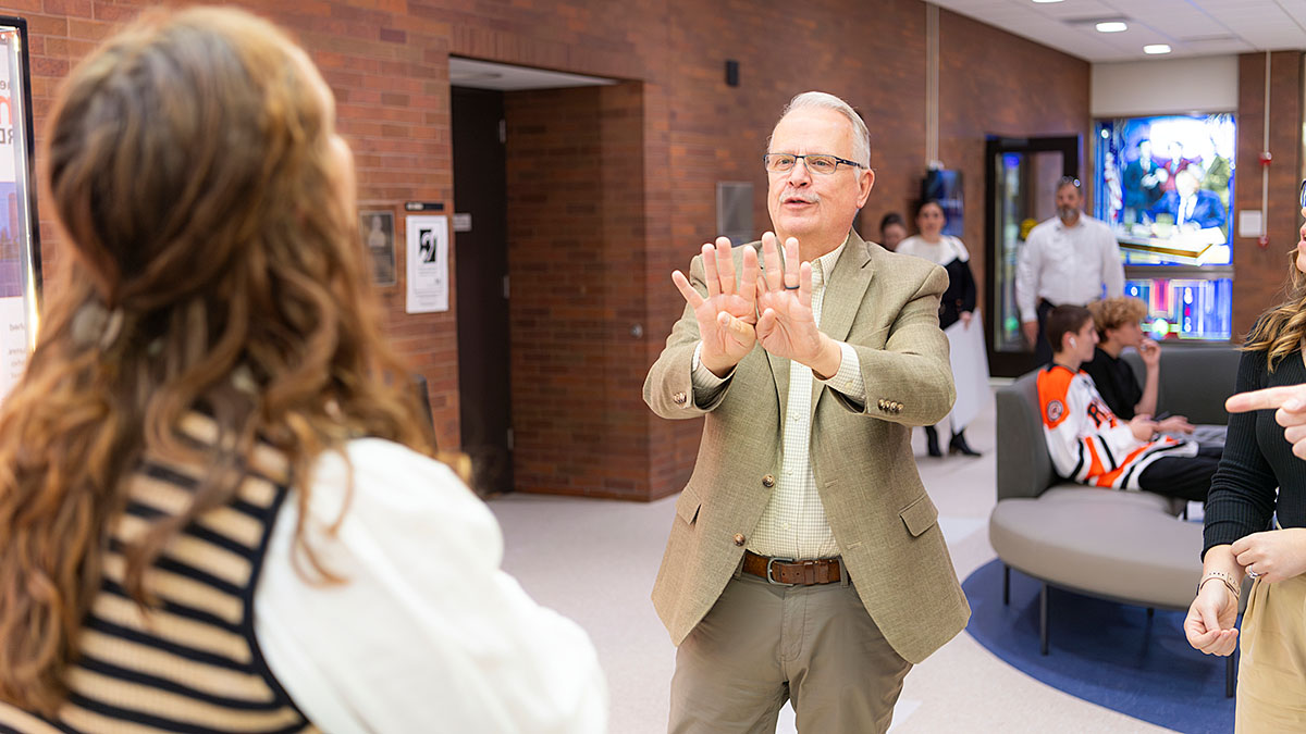 Person in beige suit jacket signing in a hallway while another person faces them; several other people are present in the background.