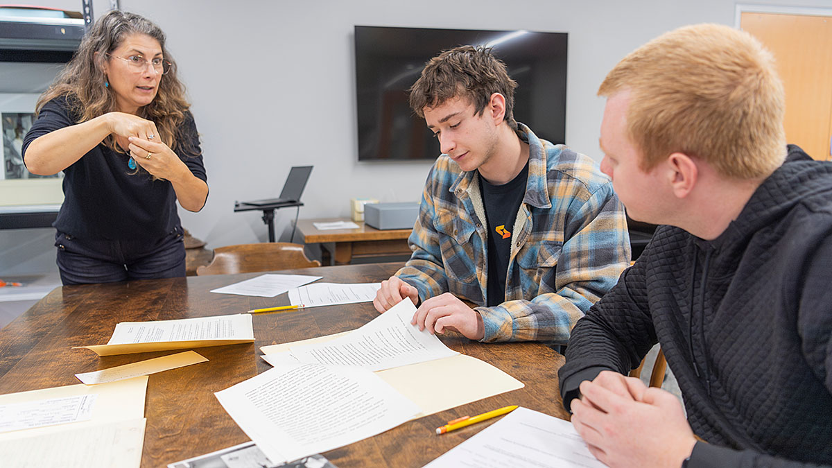 Three people are gathered around a wooden table with various papers, documents, and pencils spread out. One person is standing and signing, while the other two are seated and looking at the papers. A large screen is mounted on the wall in the background.