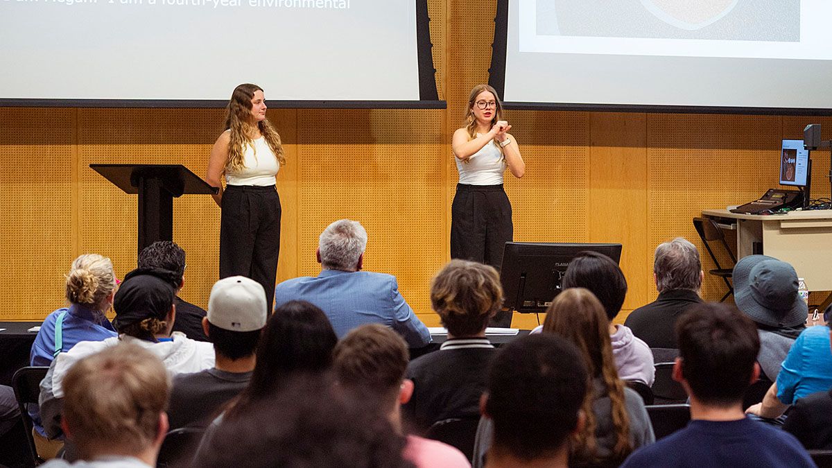 Two presenters stand at the front of a lecture hall giving a presentation to an audience seated in rows, with slides displayed on large screens behind them.