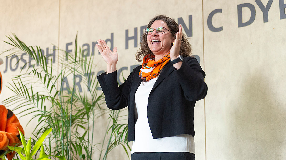 A person with curly hair and glasses, wearing a black blazer and orange scarf, smiling with raised hands in front of a wall with partially visible text and green plants in the background.