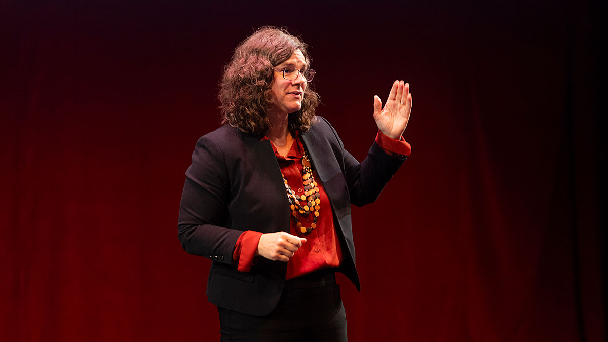 A person with curly hair, wearing glasses, a black blazer, and a red shirt, standing on stage in front of a red curtain, signing a presentation.