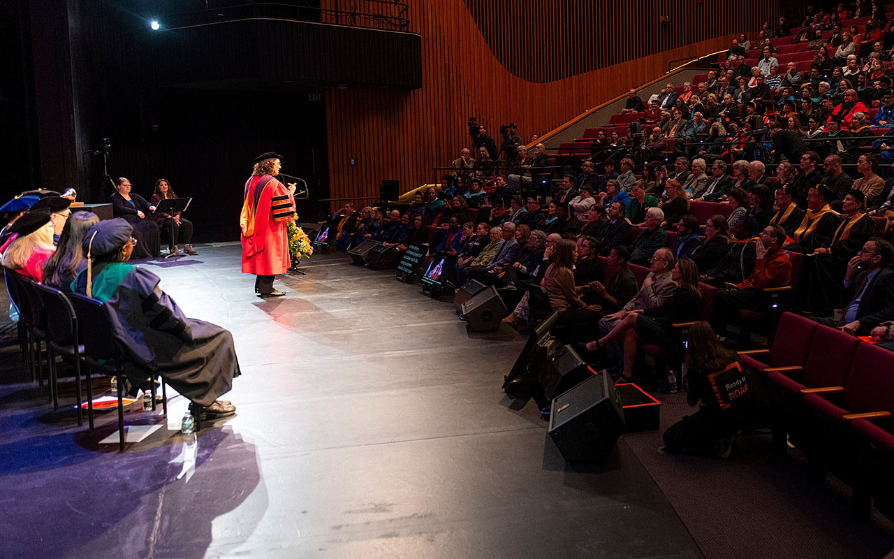 A person in academic regalia speaking on stage to a large audience in an auditorium, with several others in similar attire seated on stage.