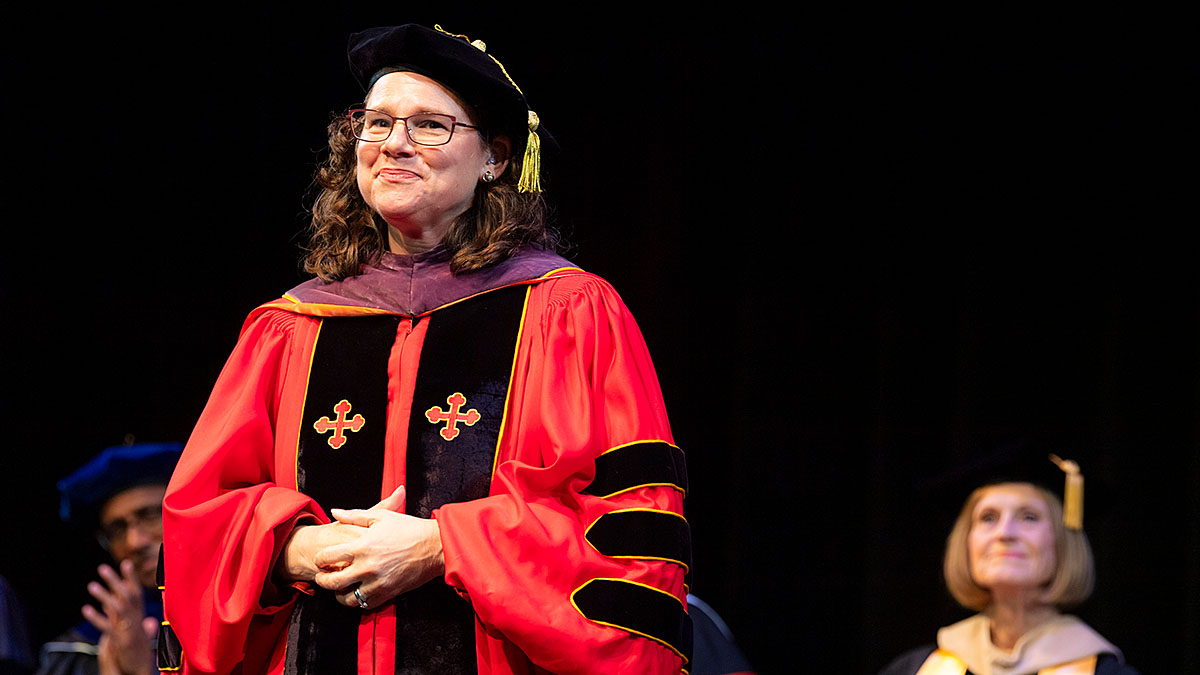 A person in academic regalia, wearing a red gown with black and gold accents and a black cap, standing on stage during a ceremony