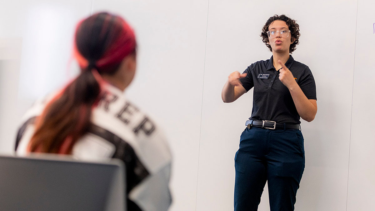 Interpreter stands at the front of a classroom while another individual watches in the foreground.
