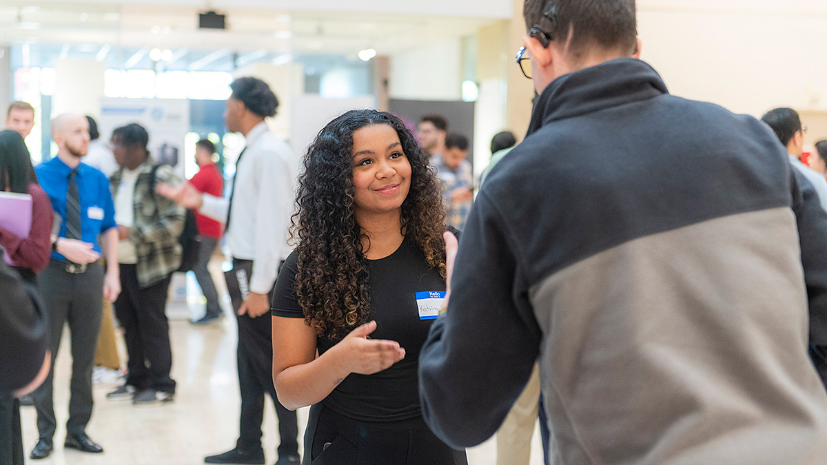 A smiling young woman with dark curly hair wearing a name tag converses with someone at a busy career fair.