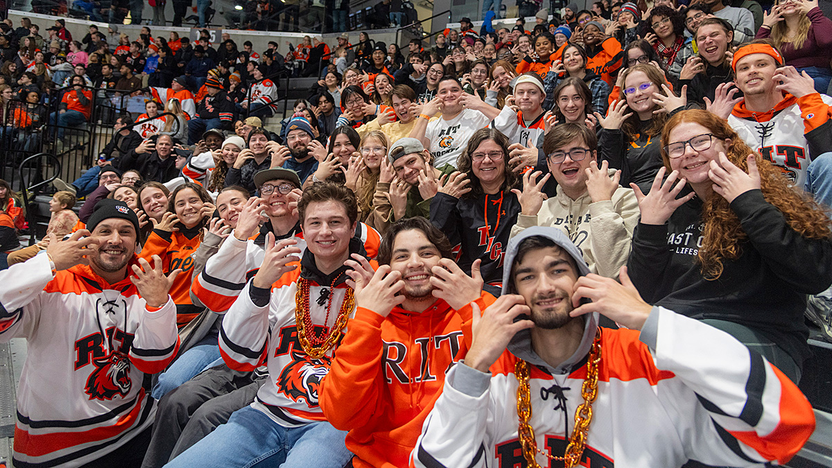 A large group of RIT (Rochester Institute of Technology) fans cheering in the stands at a hockey game, with many wearing orange and white Tigers jerseys and hoodies. Most fans are smiling and holding up both hands making the sign for tiger.