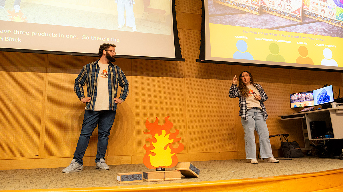 Two presenters wearing matching EmberBlock t-shirts and plaid flannels deliver a pitch on stage next to a flame cutout prop and presentation slides.