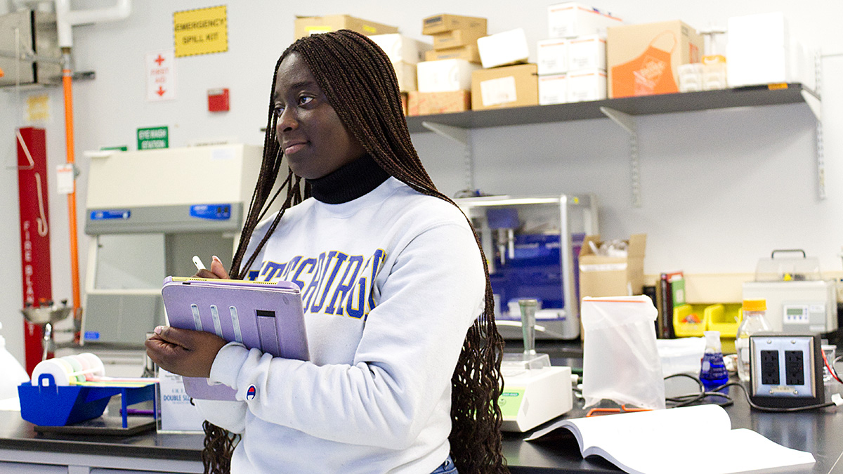 The image shows a young woman with long braided hair standing in a laboratory. She is wearing a white sweatshirt with blue and yellow lettering, and a black turtleneck underneath. She holds a tablet and a stylus in her hands and appears to be observing something or taking notes. The lab is equipped with various scientific instruments, shelves filled with boxes and supplies, and safety signs.