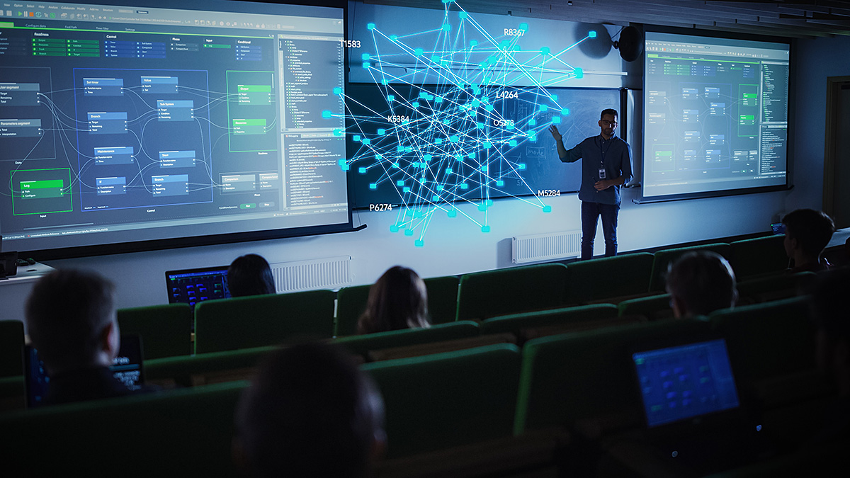 A presenter in a darkened lecture hall gestures toward a glowing blue, 3D holographic network graph floating in mid-air. Large projection screens behind him display complex software flowcharts, while silhouetted students watch.