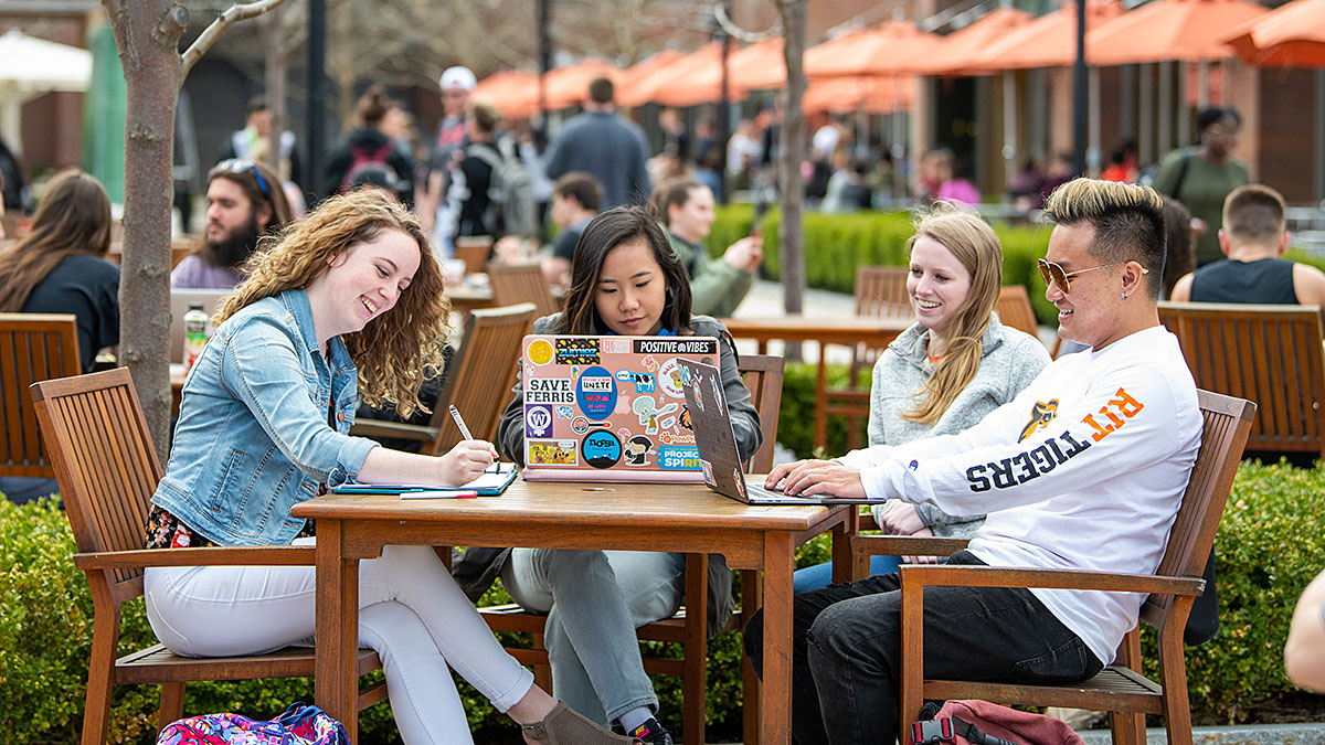 Four students site outside around a table at a busy campus location