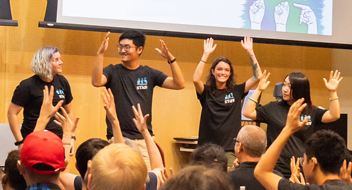 Students and instructors in auditorium with their hands up signing