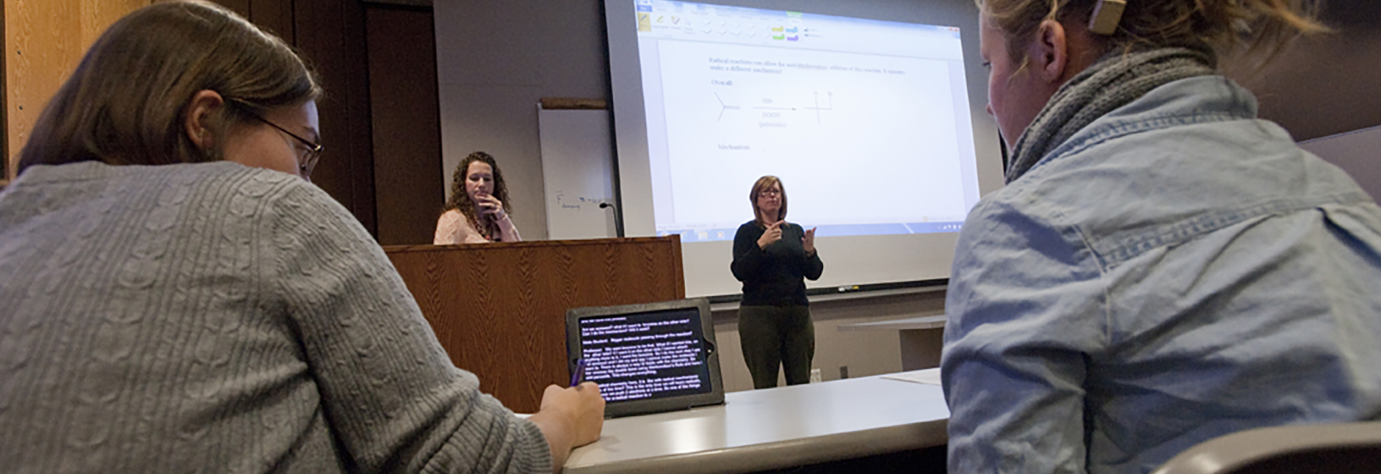 Photo of students in classroom with cprint captions assistance on monitor