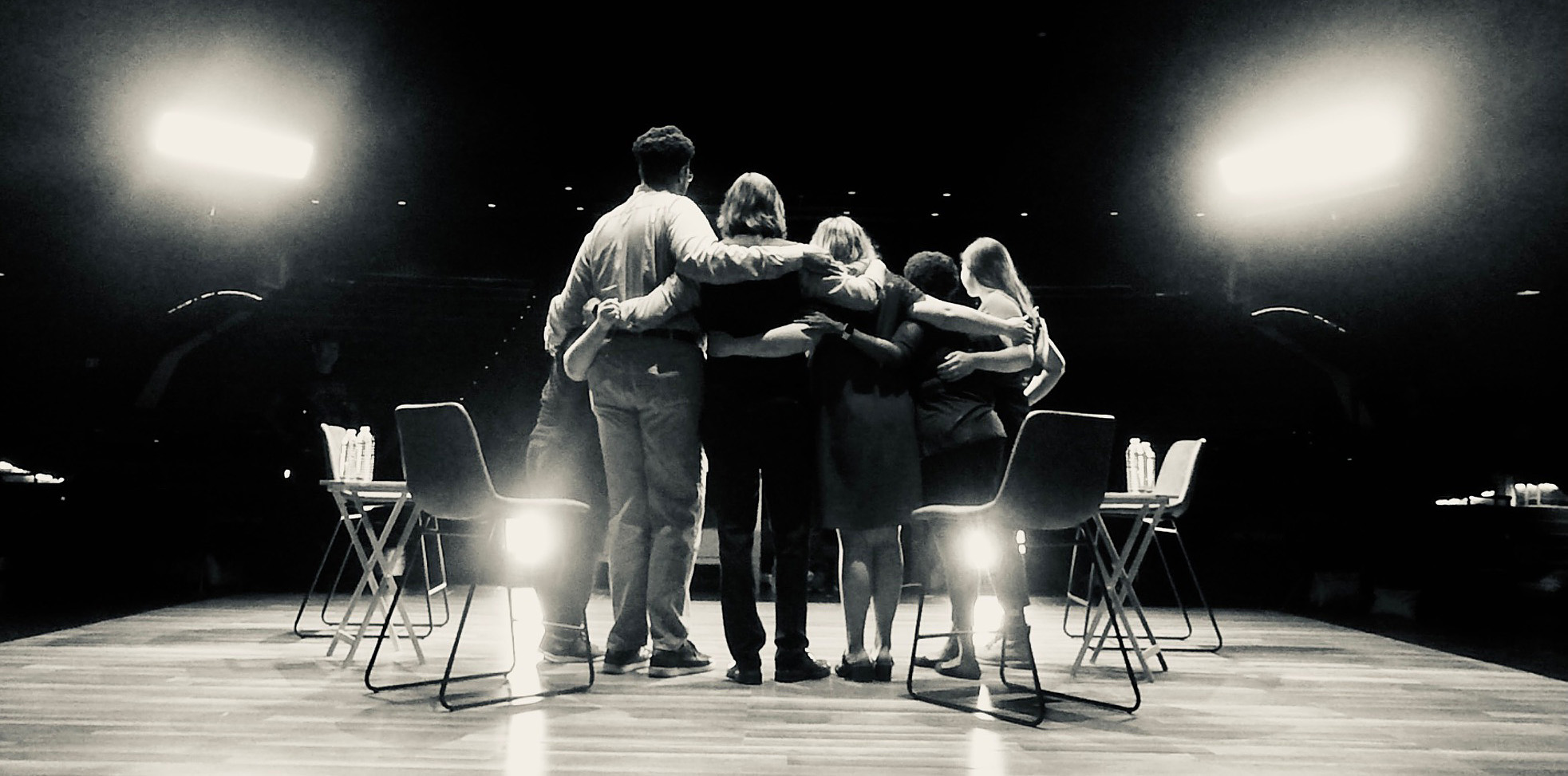 Black and white photograph of 5 people standing on stage with their arms around each other. The photo is taken behind the people, looking out at a theater.