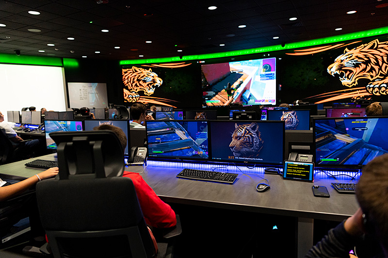 The image depicts a modern esports or gaming lab filled with students seated at gaming stations. The room is dimly lit, with blue and green accent lighting along the desks and ceiling. Each station is equipped with a high-performance gaming PC, dual monitors, keyboards, and mice. The screens display a competitive multiplayer game in progress.