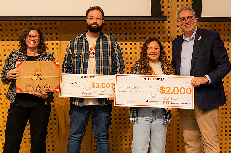 Four people pose on stage holding an EmberBlock product box and two large ceremonial checks for $3,000 and $2,000 from the Next Big Idea competition.