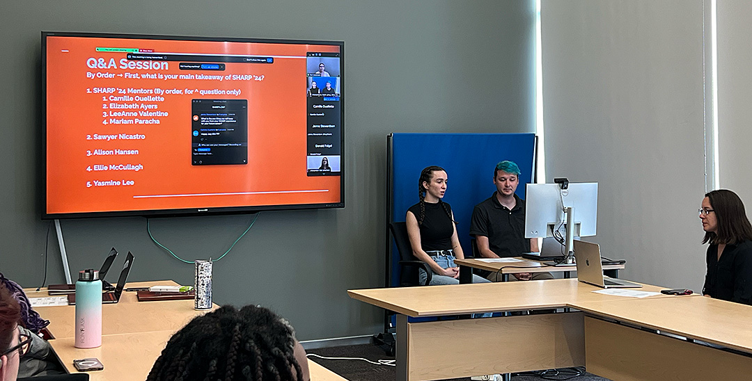 The image shows a classroom or meeting room setting with several people present. There is a large screen at the front displaying a slide.