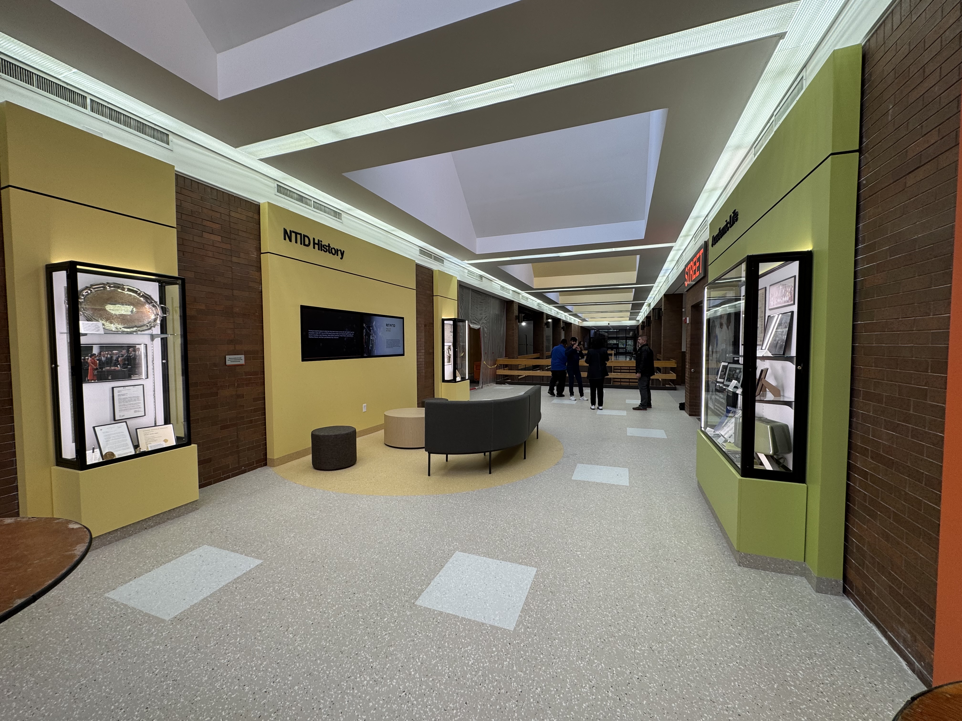 The image depicts a well-lit hallway or lobby area at the National Technical Institute for the Deaf (NTID), part of Rochester Institute of Technology (RIT). The space, known as "The Street," features modern design elements with a combination of brick walls, yellow and green accent panels, and display cases showcasing historical and technological exhibits.