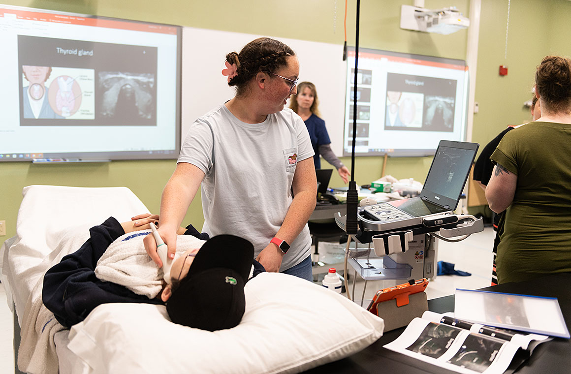 Student performing an ultrasound on a patient lying on a bed in a classroom setting, with educational slides about the thyroid gland displayed on screens in the background.
