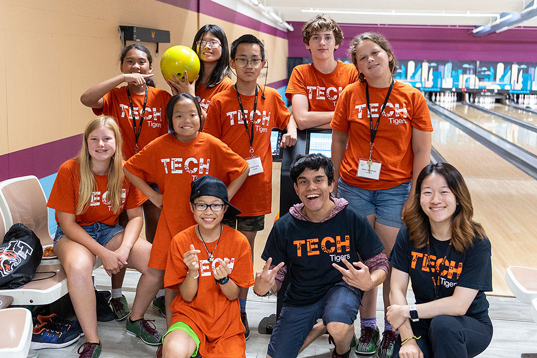 A group of ten people wearing orange and black 'TECH Tigers' t-shirts posing together in a bowling alley.