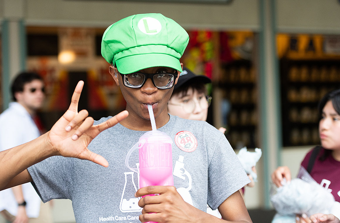 Person wearing a green hat with an 'L' emblem, holding a pink drink, and making the sign for 'love'.