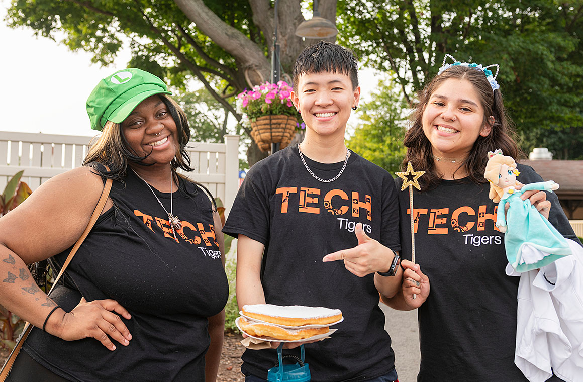 Three individuals in black 'TECH Tigers' t-shirts standing outdoors among trees and flowers, holding a stack of pancakes, a wand, and a doll.
