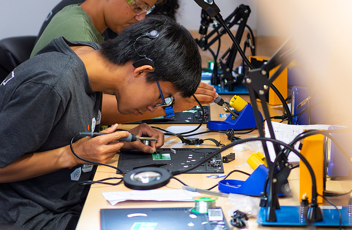 A person soldering electronic components at a workstation with various tools and equipment.