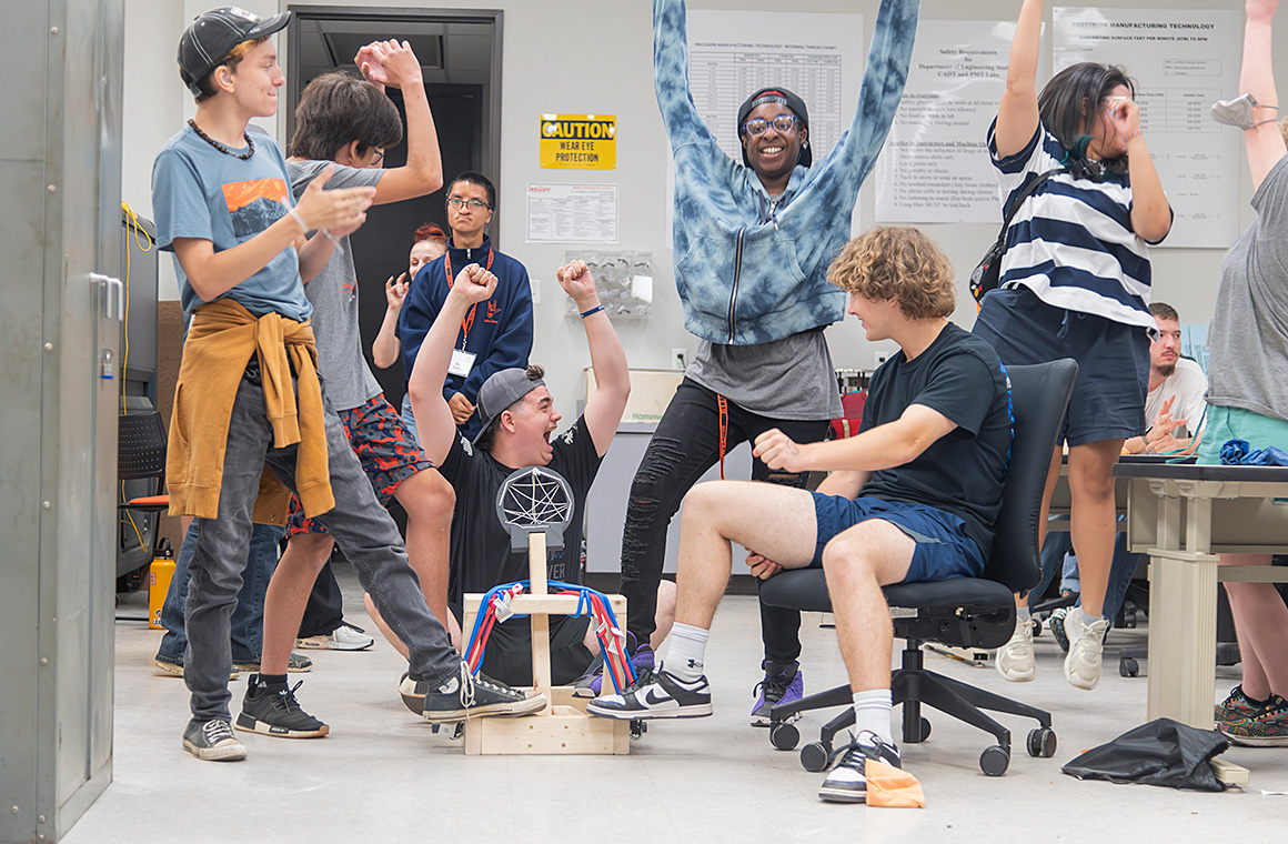 A group of students celebrating around a small wooden structure with colorful ribbons in a classroom or workshop setting