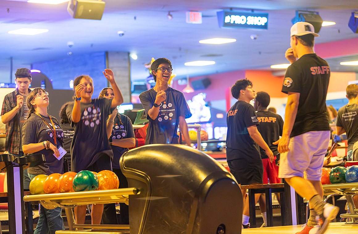A group of people at a bowling alley, with colorful bowling balls on a return rack in the foreground. Some individuals are chatting or preparing to bowl, while others walk around. The background features bowling lanes, seating, and a 'Pro Shop' sign. Several people wear shirts labeled 'STAFF.'